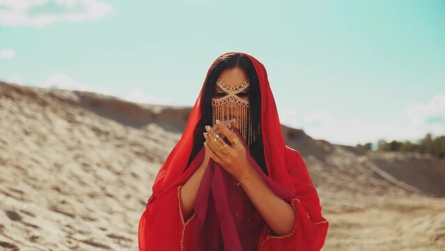 fantasy woman in desert fashion model posing dansing sexy eyes hands holding scarf golden gold metal mask veil hides face. lady queen red dress girl arab style. summer nature sand sun light blue sky