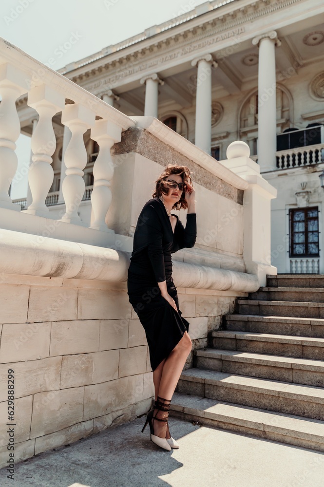 Wall mural Stylish woman in the city. Fashion photo of a beautiful model in an elegant black dress posing against the backdrop of a building on a city street