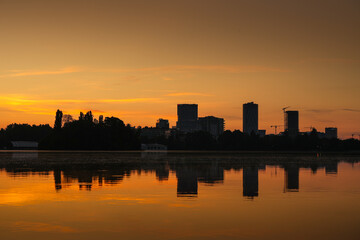 Fototapeta premium Sunrise over Bucharest. Sunrise photo in the morning dusk with orange and blue sky with the skyline of Bucharest and reflection in Herastrau Lake. Travel to Romania.