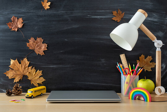 Educational tools composition. Side view photo of color pencils, plasticine, laptop, apple, lamp, bus toy, pine cones, maple leaves, stationery holder on desk with chalkboard backdrop. Perfect for ads