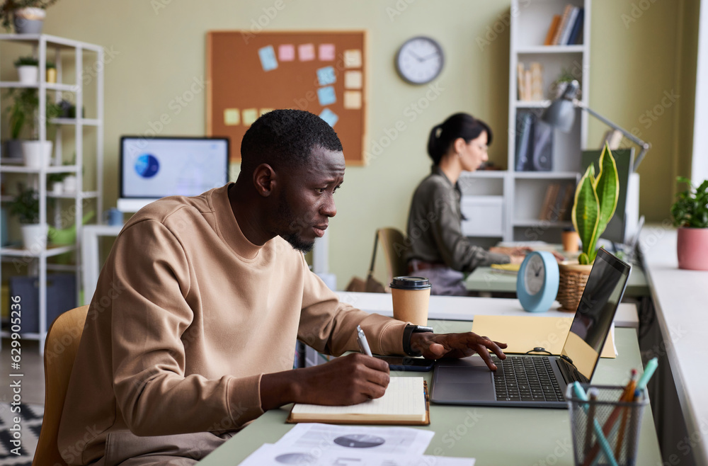 Aufkleber side view portrait of black young man working at desk in office and taking notes writing in notebook