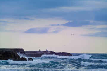 Sunset at Muriwai beach. Stormy waves splashing against the rocks where fishermen bracing the elements and seaguls circling above. Auckland, New Zealand