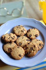 Plate of chocolate chip cookies, stack of books, reading glasses, orange soda, mobile phone and pen on the table. Hygge at home. Pastel colors, selective focus.