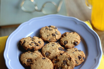 Plate of chocolate chip cookies, stack of books, reading glasses, orange soda, mobile phone and pen on the table. Hygge at home. Pastel colors, selective focus.
