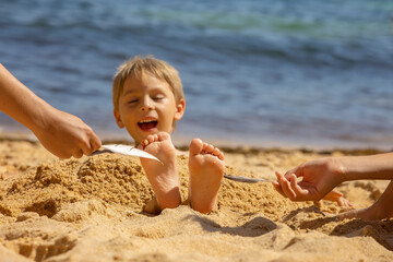 Child, tickling sibling on the beach on the feet with feather, kid cover in sand, smiling, laughing