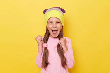 Extremely happy cheerful little girl with ponytails wearing pink shirt and beanie hat posing...