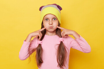 Funny little girl with ponytails wearing pink shirt and beanie hat posing isolated over yellow background standing blowing cheeks pointing with fingers