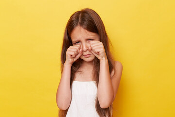 Unhappy sad little girl with long hair standing isolated over yellow background crying being...