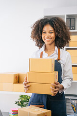 African American female holding parcels and looking to camera, standing among several boxes in modern office.