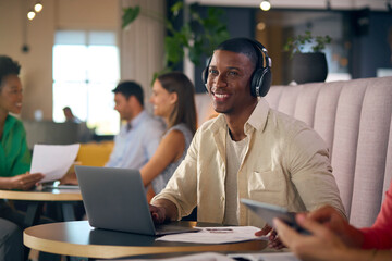 Fototapeta na wymiar Businessman Wearing Wireless Headphones Working On Laptop In Informal Seating Area Of Modern Office