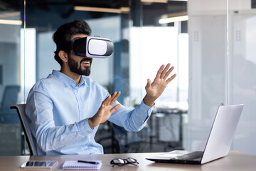 Young Indian male student sitting at a desk in the office wearing a virtual mask, watching online videos, studying, talking, gesturing with his hands