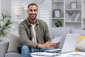 Portrait of a young Hispanic man looking at the camera with a smile, working remotely from home with a notebook and documents