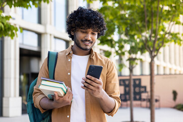 Young Indian male student using mobile phone, standing outside on street, holding books in hand, reading message, looking for campus, waiting for meeting