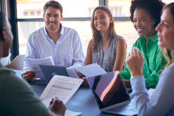 Multi-Cultural Business Team Meeting Around Office Boardroom Table With Laptops Discussing Documents