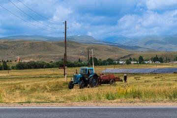Tracktor collecting hay in a mountain meadow at springtime. Tractor mowing the grass with beautiful high mountains in the background, village ecological agriculture.
