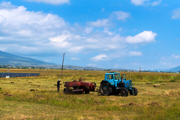 Tracktor collecting hay in a mountain meadow at springtime. Tractor mowing the grass with beautiful high mountains in the background, village ecological agriculture.
