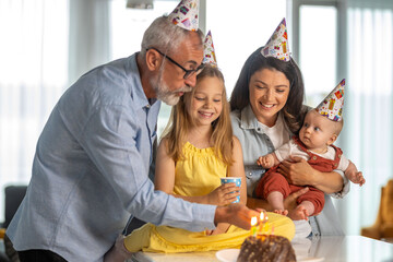 Grandfather lights the candles on the cake while granddaughter looks on and laughs with mom and her sister