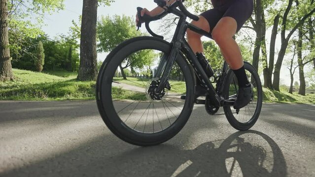 Cropped view of male cycler pedaling along paved driveway on racing bike using sunny day in city park. Efficient sports person in tight-fitting clothes conducting slow recreational trip outdoors.