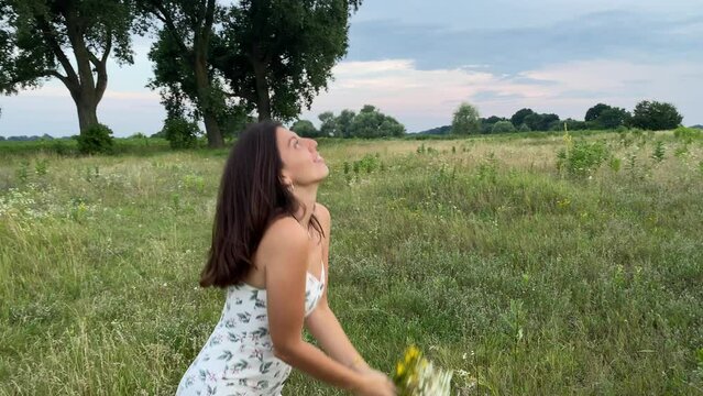 Multiracial young woman in dress enjoys leisure time at picturesque meadow in twilight. Pretty girl inhales fragrant aroma wild herbs, tosses bouquet of wildflowers. Carefree pastime in nature beauty