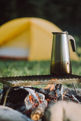 Selective focus of coffee prepared in geyser coffee maker on campfire at summer morning. Breakfast drink while hiking. Bonfire on background tent and forest. Concept adventure active vacations outdoor
