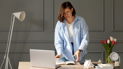 Beautiful young businesswoman which is wearing a white t-shirt, blue shirt and light jeans is standing near a desk with a laptop and work supplies on it flips through the notebook