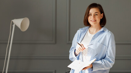 Business girl wearing a white t-shirt, blue shirt and light jeans, is standing on a gray background, holding a pen and glasses in one hand