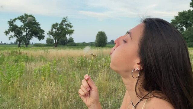 Multiracial young woman blows all dandelion seeds at once, making dandelion wish come true. Carefree profile portrait on backdrop serene countryside and cloudy sky. Nature beauty in picturesque meadow