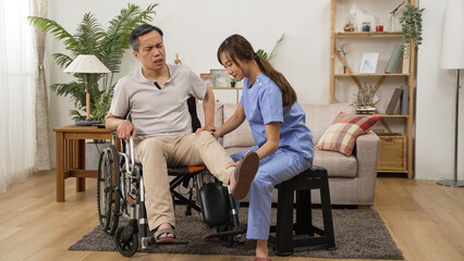 asian female care attendant helping senior man on wheelchair doing rehab training. she slowly lifts his injured leg while he shows painful face in the living room at home