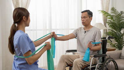 selective focus of asian senior disabled man strengthening his bicep by doing physiotherapy resistance training using elastic band. he sits on wheelchair with his care attendant at home
