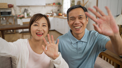 mobile cam view of cheerful asian Taiwanese adult daughter and father waving hi to camera and greeting their friends with smile while having online video chat at home