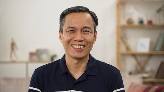 shoulder shot portrait of happy asian senior man looking at the camera with a big smile against modern cozy home background