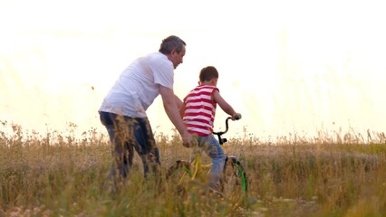 Father in white shirt supports bike of son learning to ride at countryside