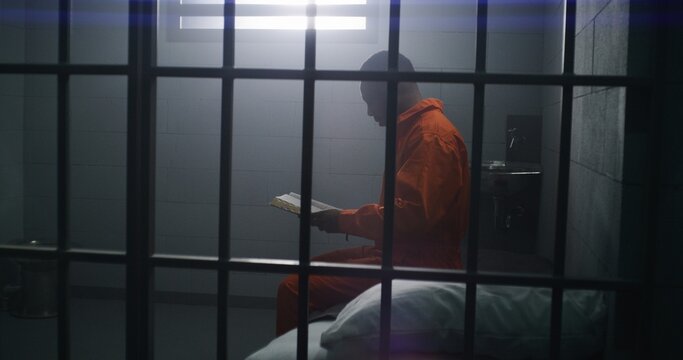 African American prisoner in orange uniform sits on the bed, reads Bible in prison cell. Male criminal serves imprisonment term for crime in jail or detention center. Faith in God. View through bars.