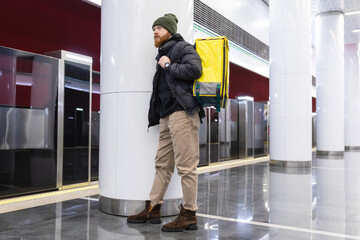 A stylishly dressed delivery man with a yellow backpack is waiting for a subway car. Food delivery in a big city