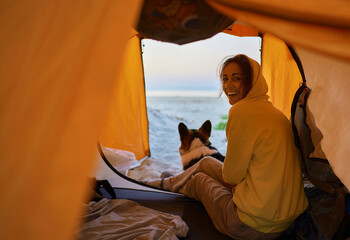 Happy young millennial girl with her puppy enjoying camping weekend and sitting inside orange tent....
