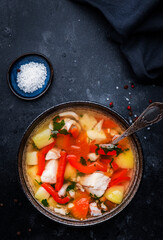 Fish soup with cod, red paprika, potatoes, tomatoes and parsley in soup bowl on black table background, top view