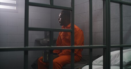 African American prisoner in orange uniform sits on the bed behind bars, reads Bible in prison cell. Criminal serves imprisonment term for crime in jail. Detention center or correctional facility.