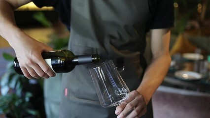 Waiter in a dark-green apron pouring red wine into wineglass. Young guy serving customers red wine in cafe. Fine dining restaurant concept.