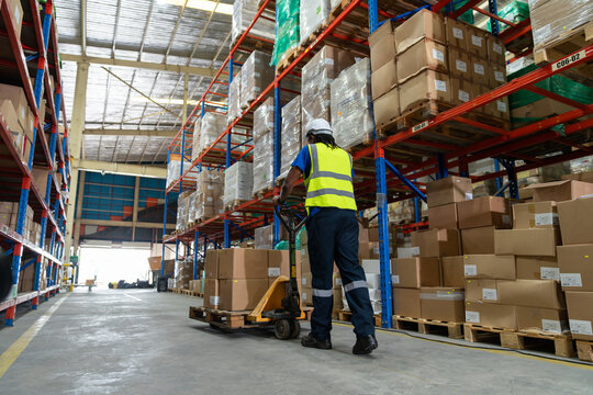 African American man worker using pallet truck for move cardboard boxes. Storehouse employee in uniform working and unloading goods in warehouse. Logistics, Distribution Center concept.