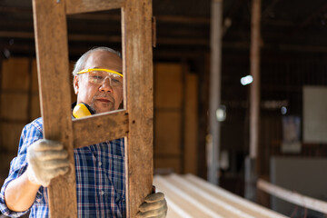 Asian senior carpenter man wearing goggles protective look at old window frame at carpentry workshop. Old male retirement renovating furniture from old wood.