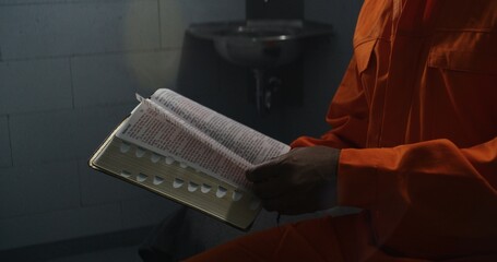 African American prisoner in orange uniform reads Bible in prison cell, turns pages. Male criminal, inmate serves imprisonment term for crime in jail or detention center. Concept of faith in God.