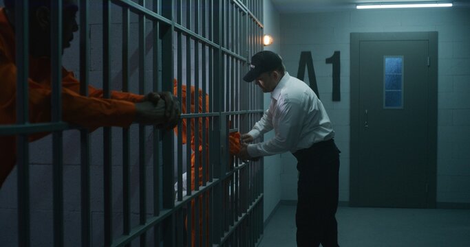 Warden brings new prisoner in jail cell and takes off his handcuffs. African American criminals serve imprisonment term in correctional facility or detention center. Guilty murderers in prison cells.