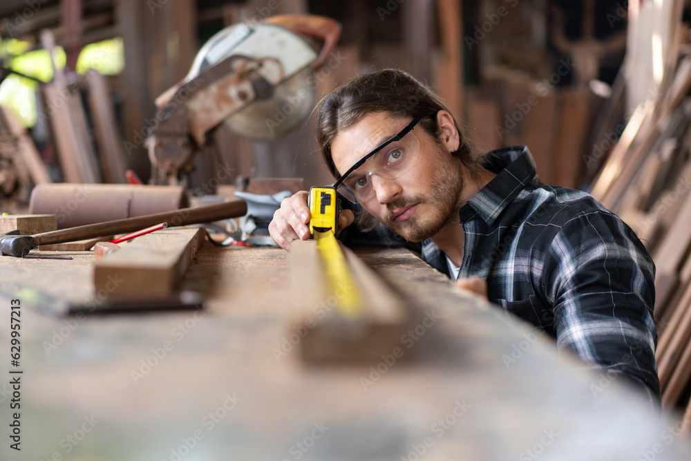 Wall mural Carpenter man in casual clothes and wearing goggles protective using ruler measuring tape wood plank materials for making furniture at carpentry workshop