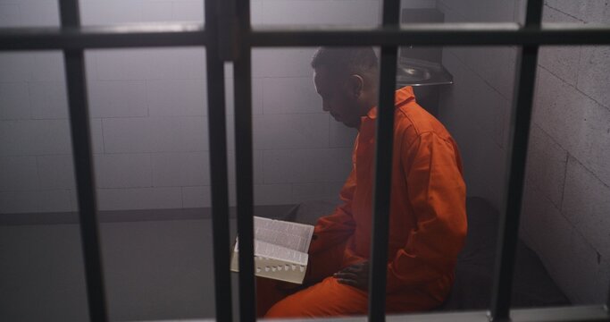 African American prisoner in orange uniform sits on bed behind bars, reads Bible in prison cell. Male criminal serves imprisonment term for crime in jail. Detention center or correctional facility.
