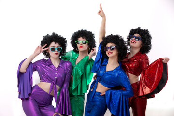 A group of girls in colorful flared suits and afro wigs pose against a white background. Disco...