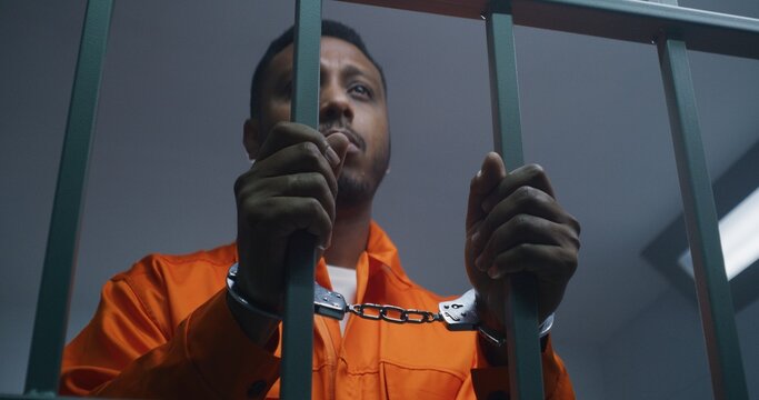African American man in orange uniform keeps hands in handcuffs on jail cell bars. Depressed murderer serves imprisonment term in prison. Guilty prisoner in correctional facility or detention center.