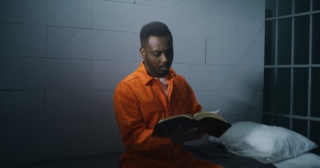 African American prisoner in orange uniform sits in prison cell, reads Bible. Male criminal serves...
