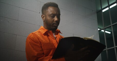 African American prisoner in orange uniform sits in prison cell, reads Bible. Male criminal serves...