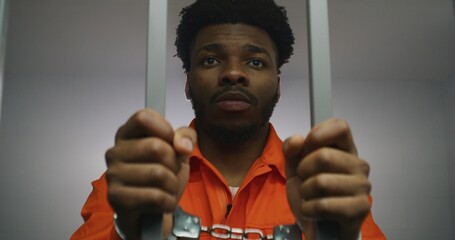African American prisoner in orange uniform keeps hands in handcuffs on jail cell bars. Guilty...