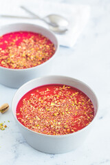 Malabi traditional Arabic dessert. Milk pudding with pink syrup and pistachio in gray bowl, white marble background.